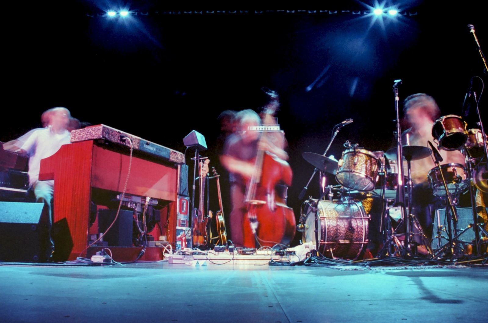 A trio plays organ, upright bass and drums on a low-angle stage, all three blurred in motion under blue lights.